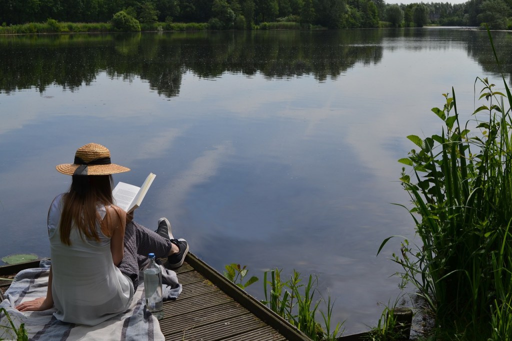 A person sitting on a wooden dock by a tranquil lake, reading a book while wearing a straw hat, with a bottle of water nearby and greenery surrounding the scene.