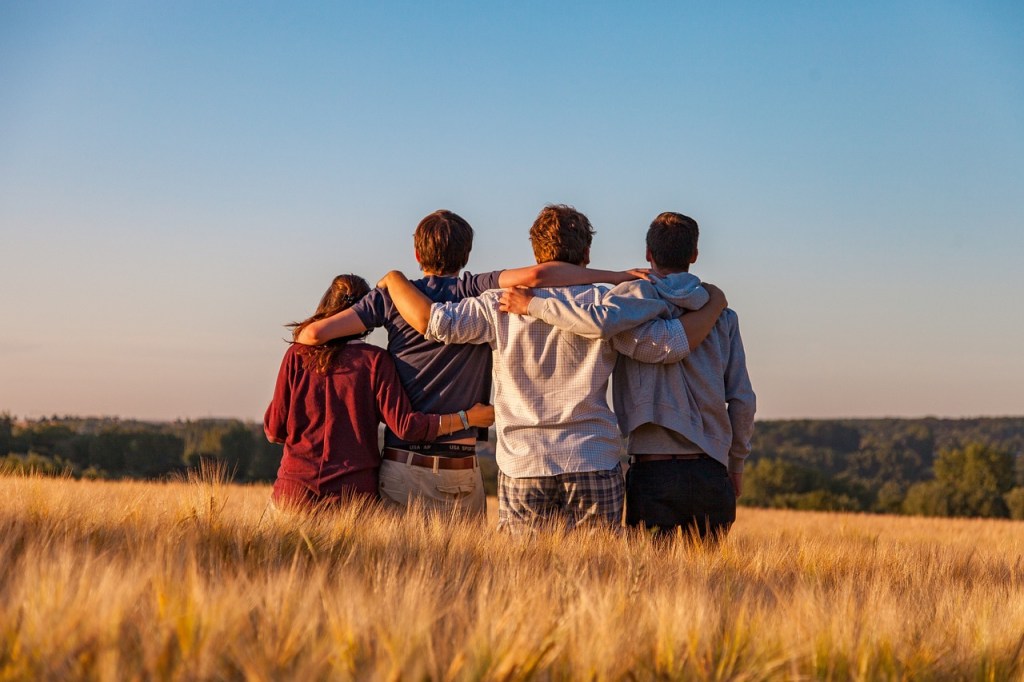 Four young adults standing together in a field, looking out towards a scenic view with arms around each other.