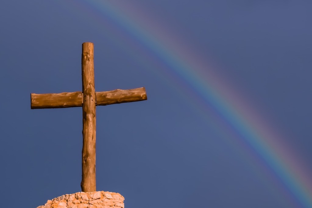 A wooden cross stands atop a rocky base against a dark sky, with a rainbow arching in the background.