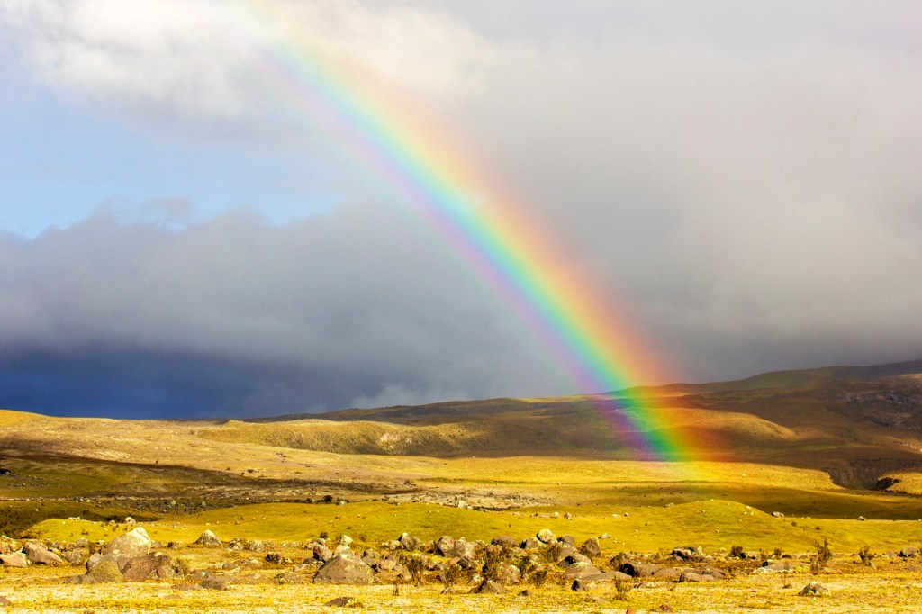 A vibrant rainbow arches across a cloudy sky, illuminating a serene landscape with rolling hills and scattered rocks.