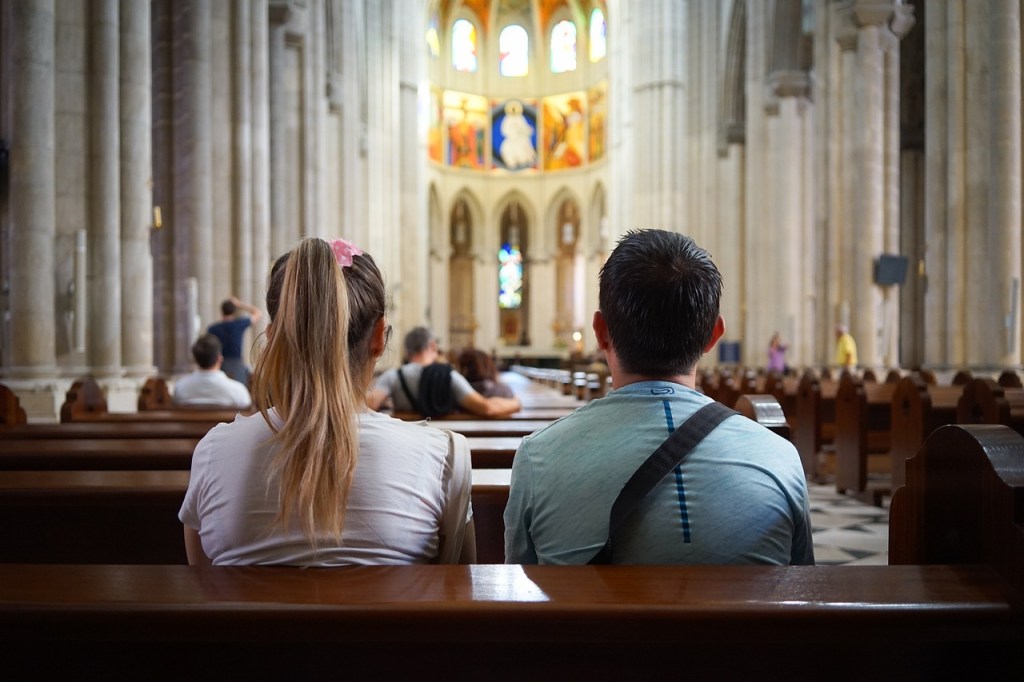 View of a church interior with two young adults sitting in wooden pews, facing the front, surrounded by other attendees.