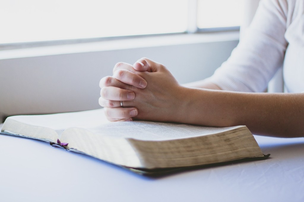 A person's hands are clasped in prayer over an open Bible, with sunlight streaming through a window in the background.