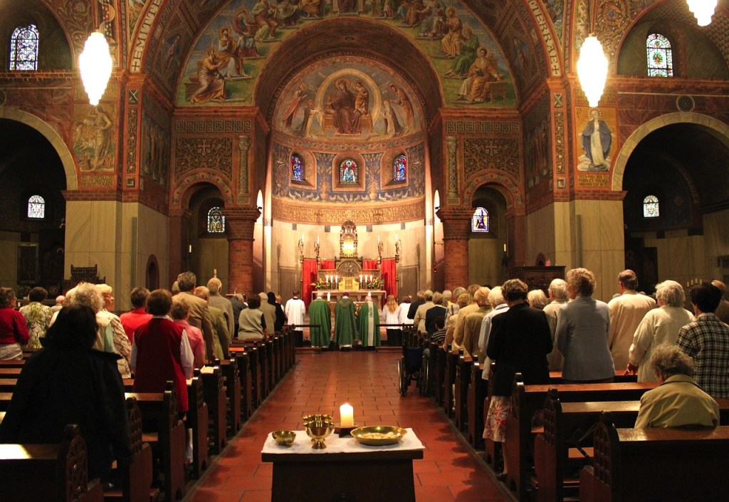 Interior of a church filled with congregants engaged in worship, featuring stained glass windows and intricate murals.