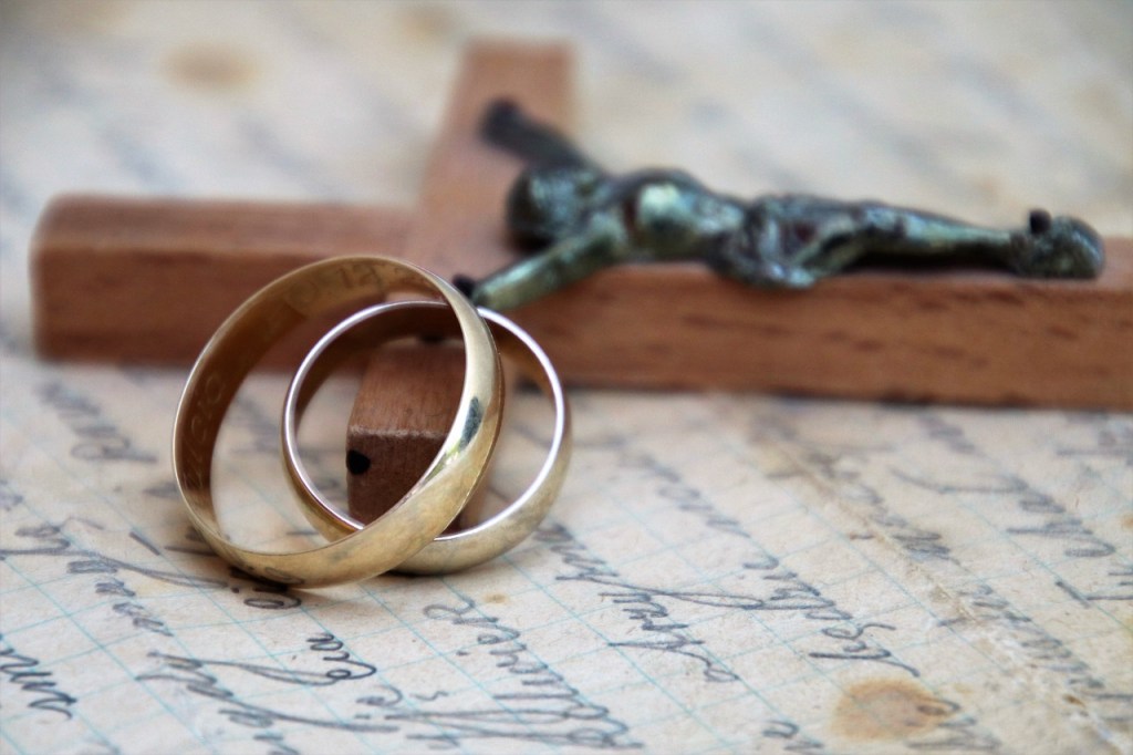Two wedding rings resting on a cross, set against a textured background of paper with handwritten notes.