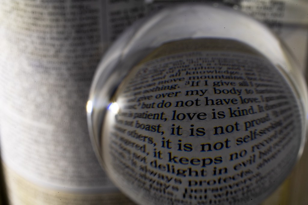 An artistic image showing a glass sphere in close-up, reflecting text from a page containing a quote about love, highlighting the phrases 'love is patient' and 'love is kind.'