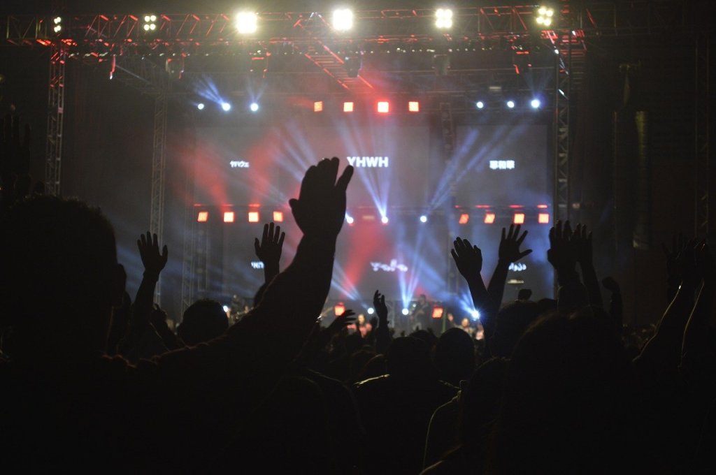 A crowd of people at a worship event with hands raised in praise, illuminated by lights and screens.