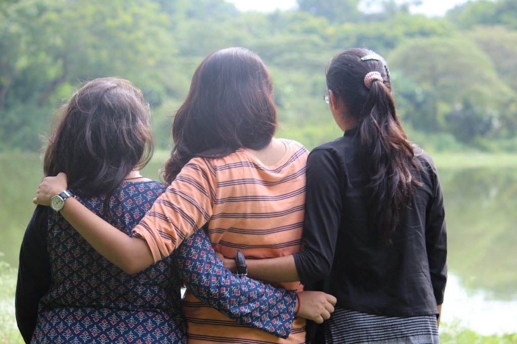 Three friends standing side by side by a lake, with arms around each other, enjoying the view.