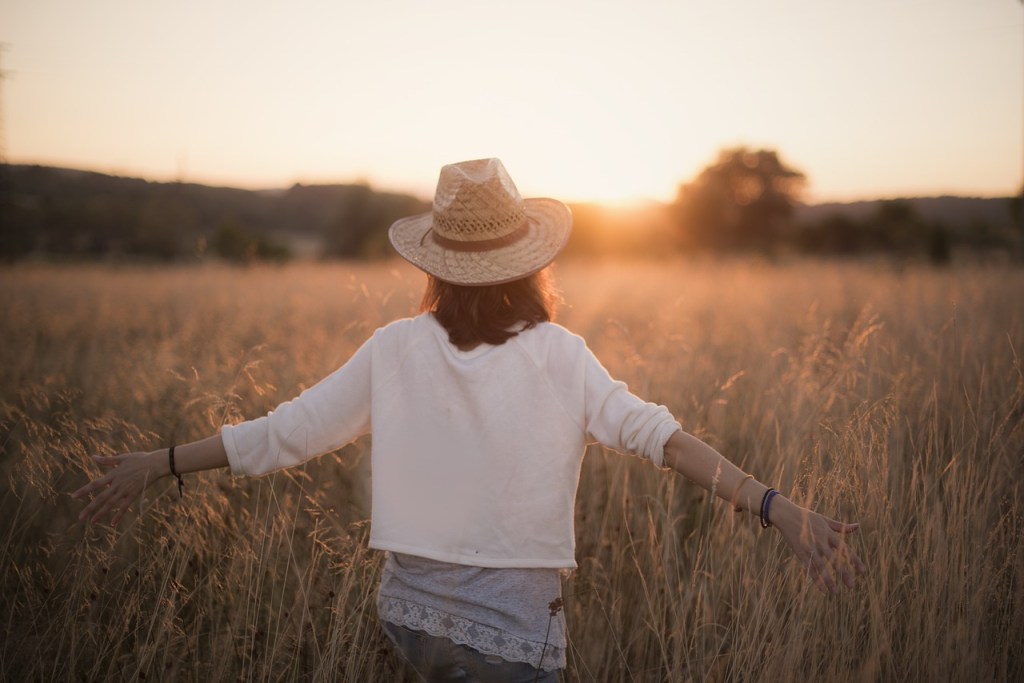 A woman with a straw hat stands in a golden field at sunset, arms outstretched, enjoying the moment.