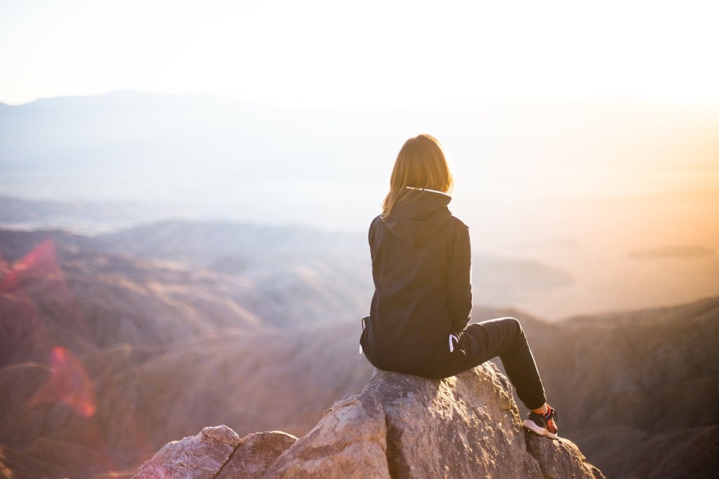 A person sitting on a rock overlooking a vast landscape during sunset, embodying a sense of reflection and solitude.