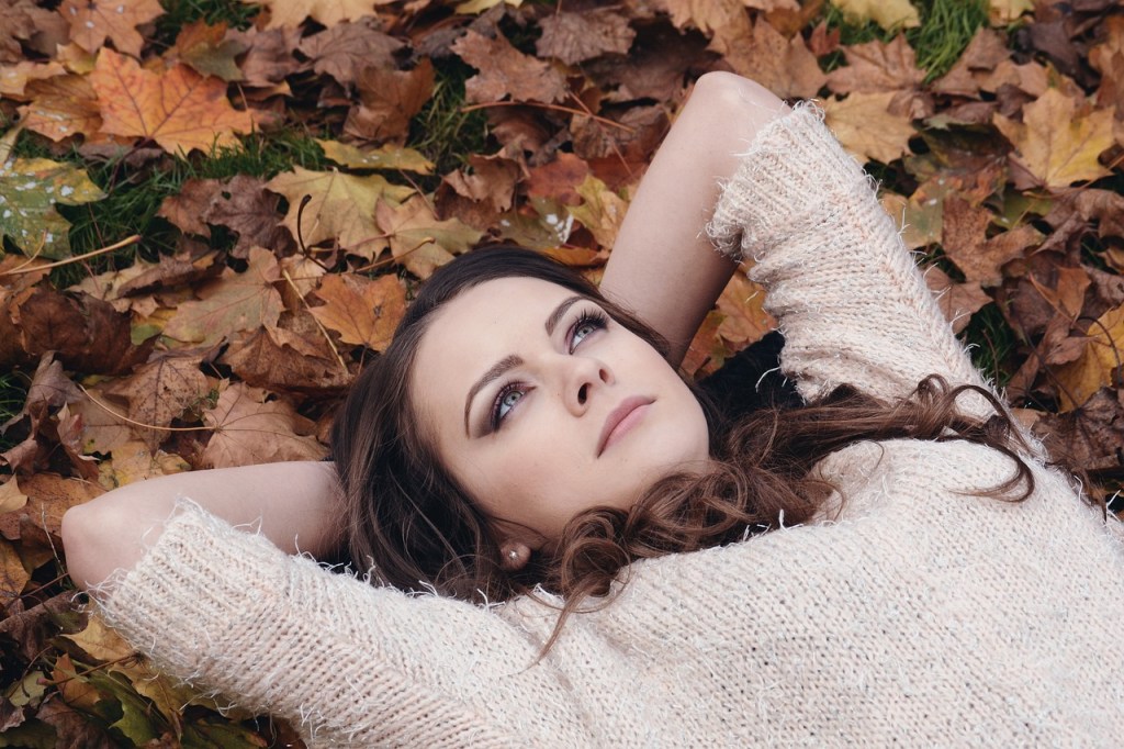 A person lying on a bed of autumn leaves, looking up thoughtfully while wearing a cozy sweater.