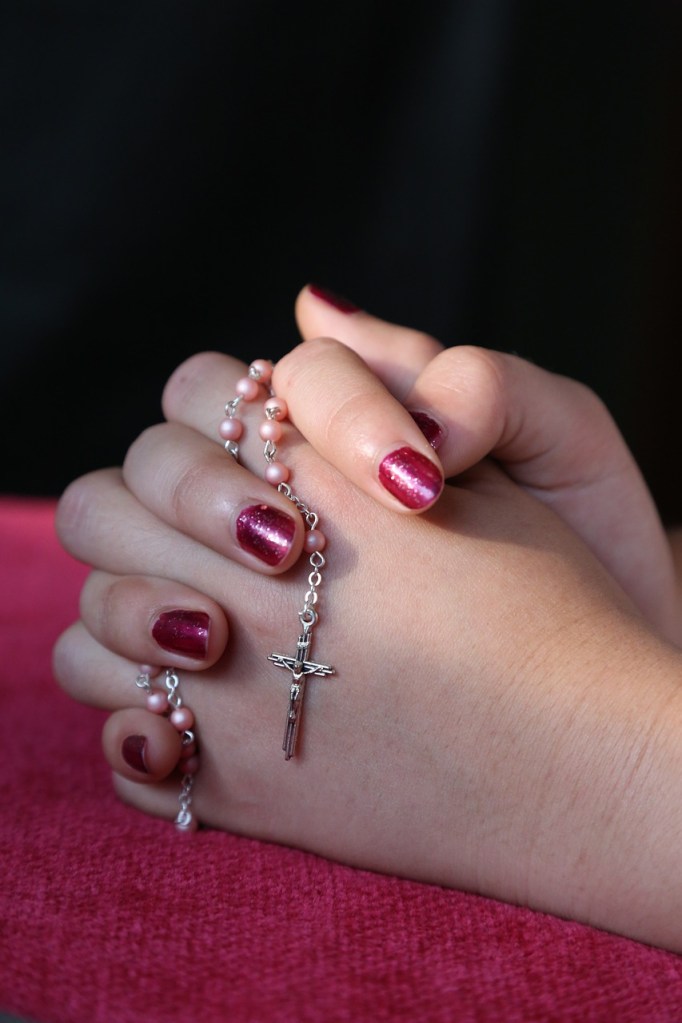 Hands clasped in prayer, adorned with a silver rosary featuring a cross, resting on a pink surface.