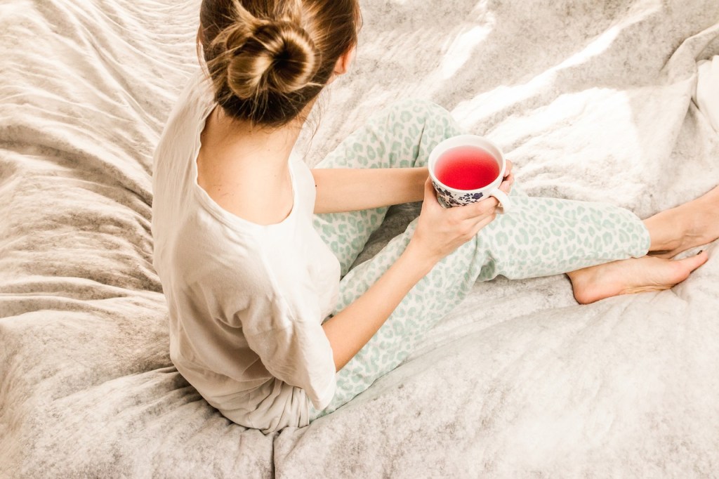 A woman sitting on a bed holding a cup of tea, wearing comfortable clothing with a relaxed posture.