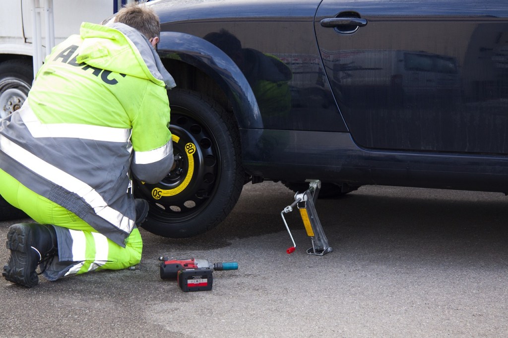 A roadside assistance worker in a bright green uniform is kneeling by a car, changing its tire with a jack and power tool.