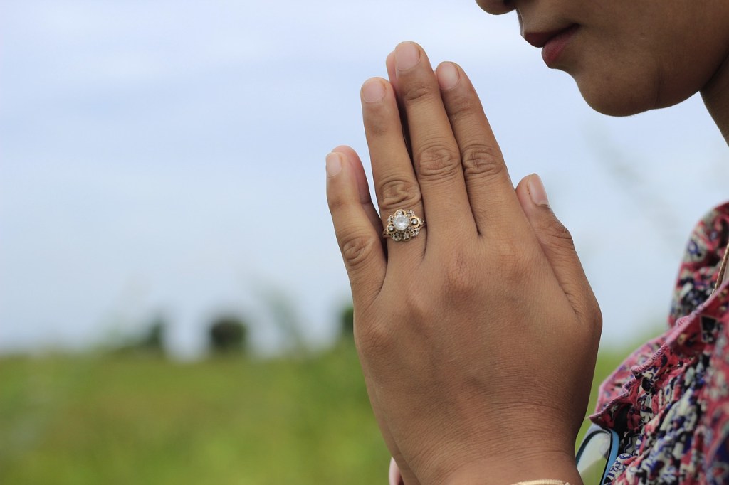 A person with clasped hands in prayer, wearing a decorative ring, with a blurred green field and cloudy sky in the background.