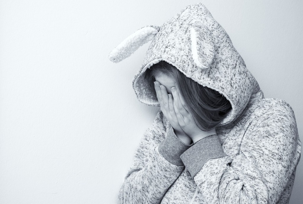 A young person wearing a gray hoodie with bunny ears is seen slightly hunched over, covering their face with their hands, conveying a sense of sadness or frustration against a plain white background.