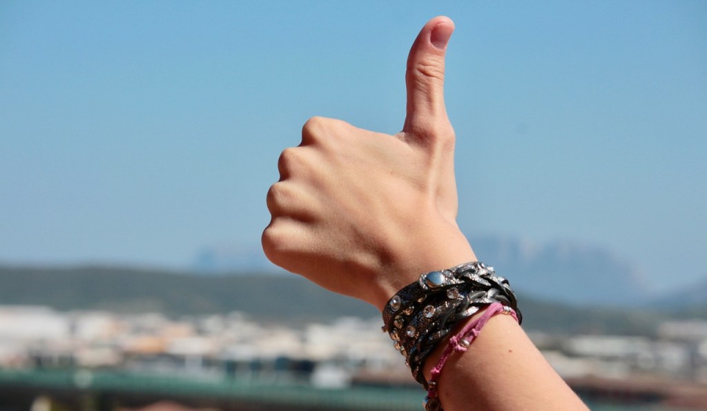 A hand with a thumb up gesture, adorned with multiple bracelets, against a clear blue sky and blurred background.