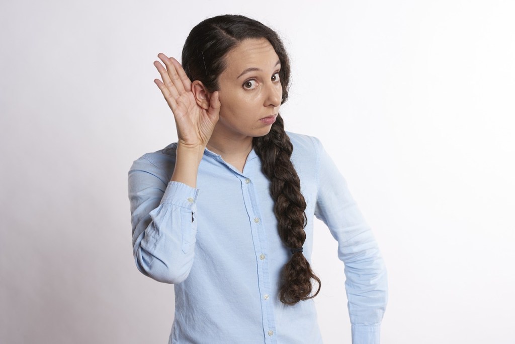 A woman with a braid wearing a light blue shirt, cupping her hand to her ear, appears to be listening intently.