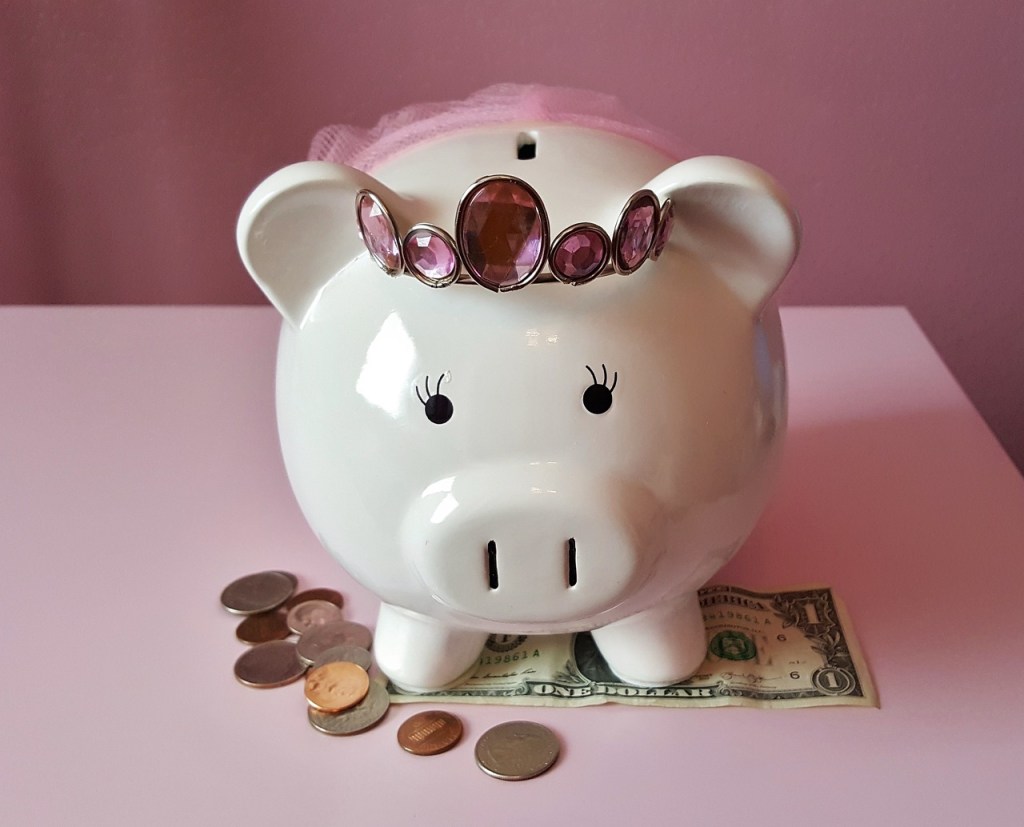 A decorative white piggy bank with a pink crown sits on a dollar bill, surrounded by various coins on a pink background.