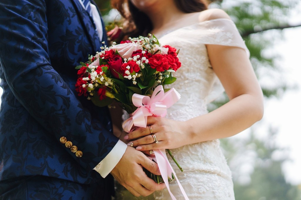 Couple holding a bouquet of red and white flowers, with the bride wearing an elegant white dress and the groom in a blue patterned suit.