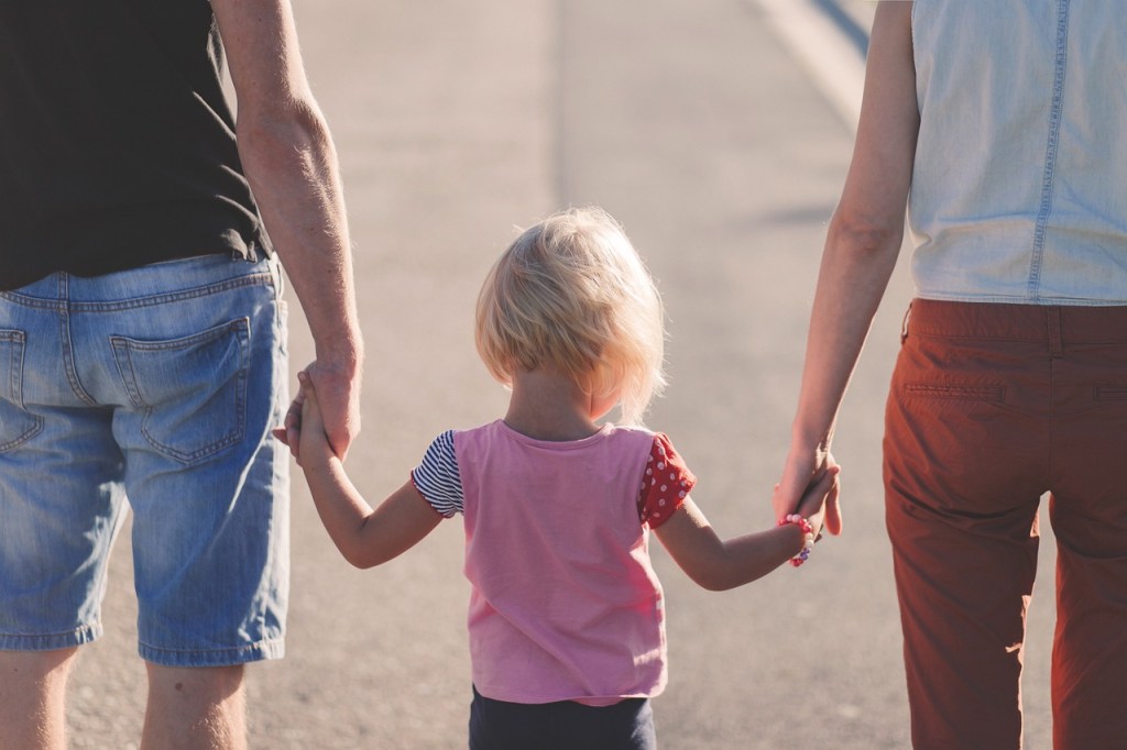 A child holding hands with two adults while walking on a sunny path.