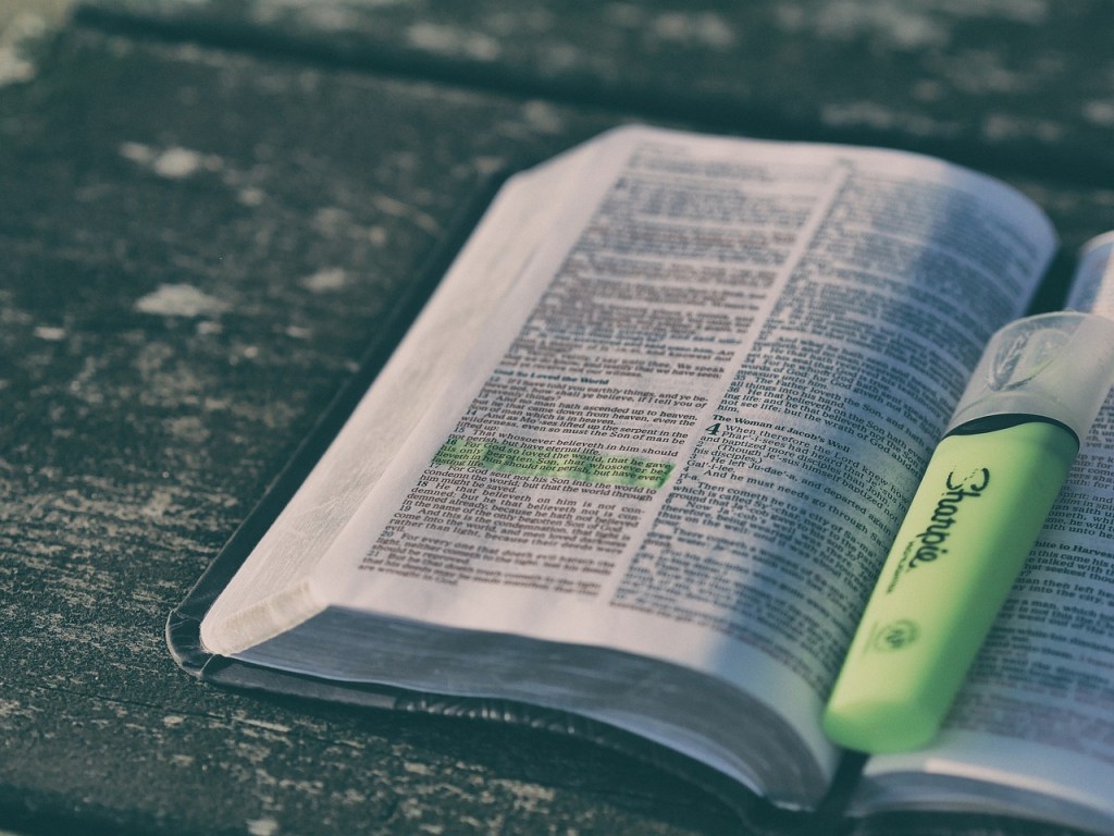 An open Bible with highlighted verses and a green highlighter on a wooden surface.