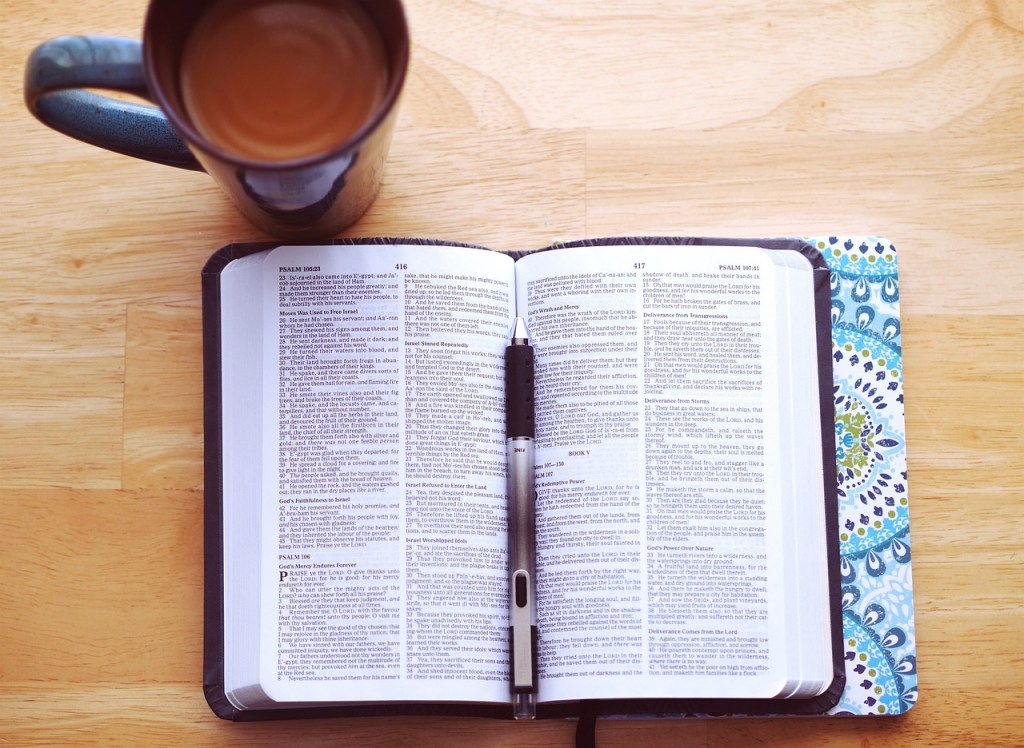 An open Bible resting on a wooden table with a black pen and a coffee mug beside it.