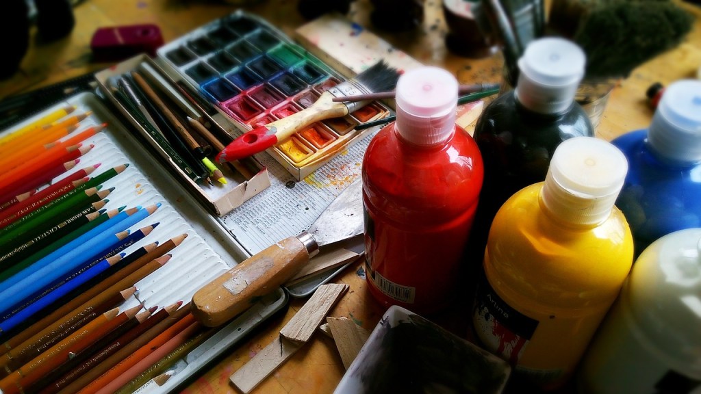 A collection of art supplies including colored pencils, paint tubes, and a watercolor palette on a wooden table.