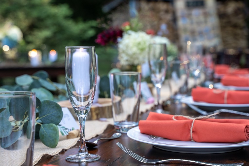 A beautifully set dining table outdoors, featuring elegant glassware and neatly arranged plates with orange napkins, surrounded by greenery and soft lighting.