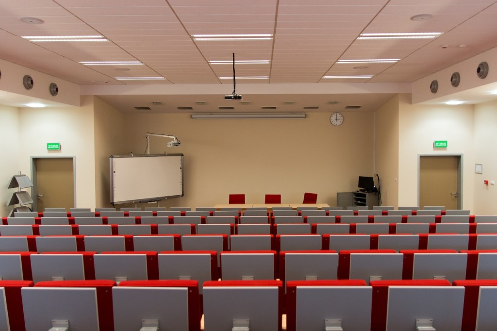 A spacious college lecture hall with rows of red and gray seating, a whiteboard at the front, and a clock on the wall.