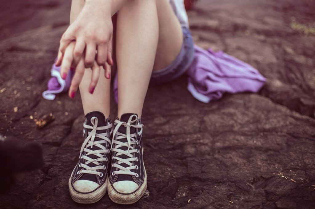 A close-up of a person's hands resting on their knees while sitting on a rocky surface, wearing black and white sneakers.