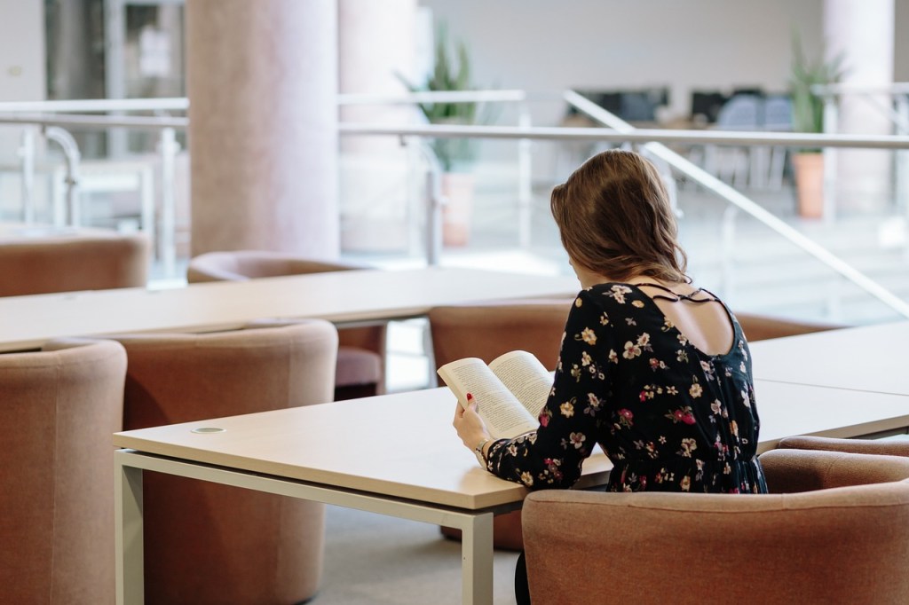 A young woman sitting at a study table in a college library, reading a book while dressed in a floral top.