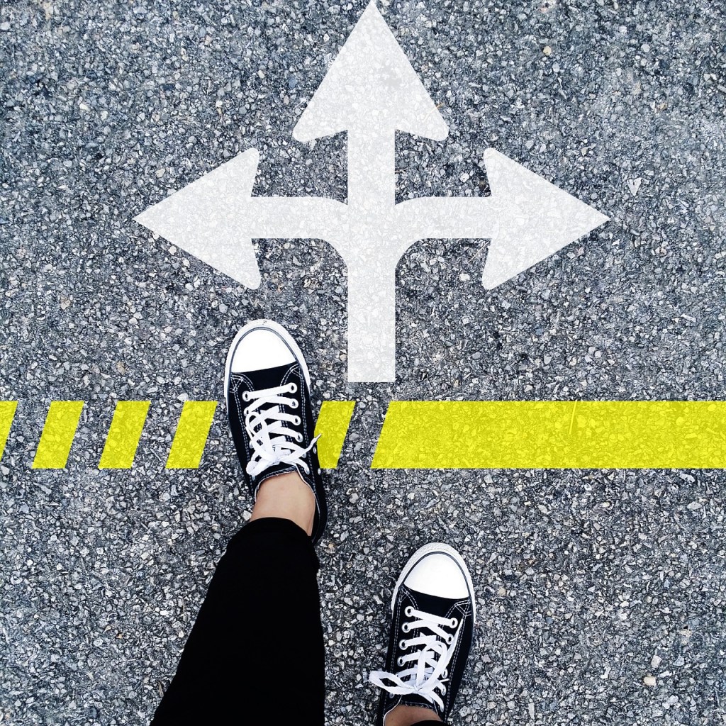A person's feet in black sneakers standing on a road marked with directional arrows and a yellow line.