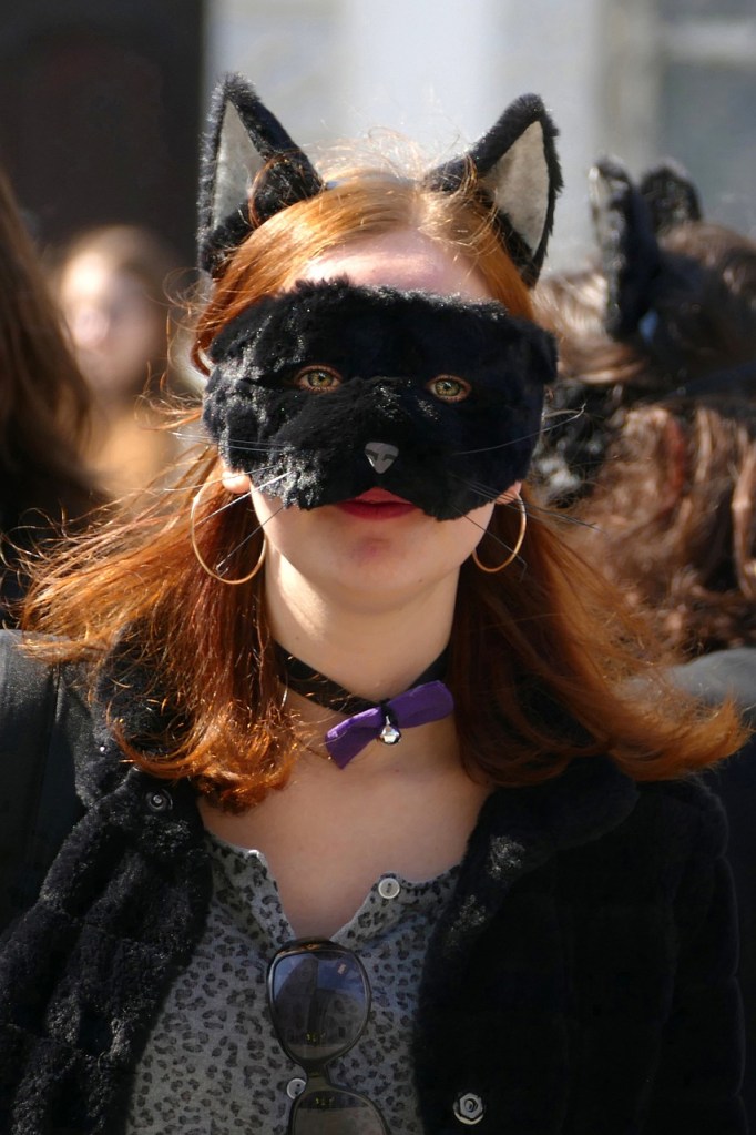 A person with red hair wearing a black cat mask and ears, with a purple choker and sunglasses hanging from their shirt. The background features blurred figures of other people.