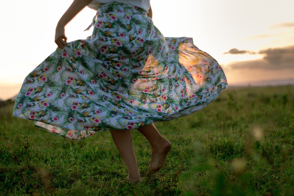 A person wearing a floral skirt twirls in a grassy field during sunset, capturing a moment of joy and freedom.