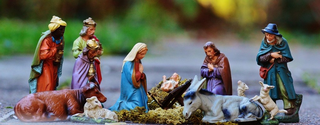 A nativity scene featuring figurines of Mary, Joseph, baby Jesus, and various shepherds and animals, set against a blurred outdoor background.