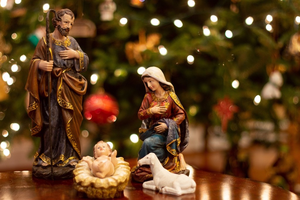 Nativity scene featuring Mary, Joseph, and baby Jesus with a white lamb, set against a backdrop of Christmas lights.