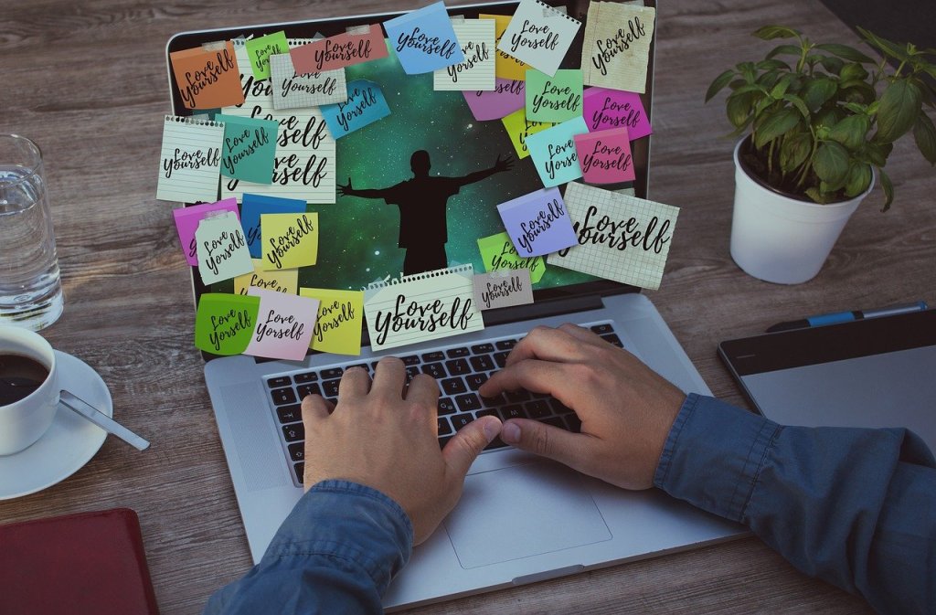 A person typing on a laptop surrounded by colorful sticky notes that read 'Love Yourself', with a plant and coffee cup nearby.