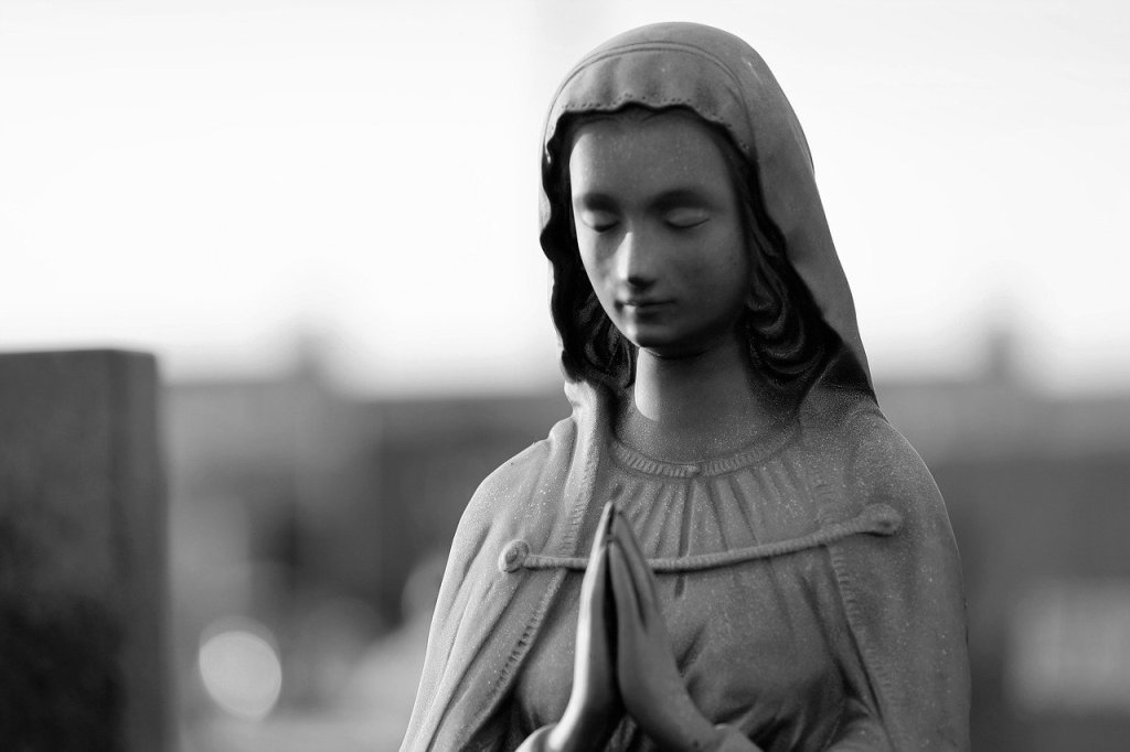 A black and white statue of Mary, mother of Jesus, with her hands clasped in prayer and her eyes closed, conveying a sense of peace and reverence.
