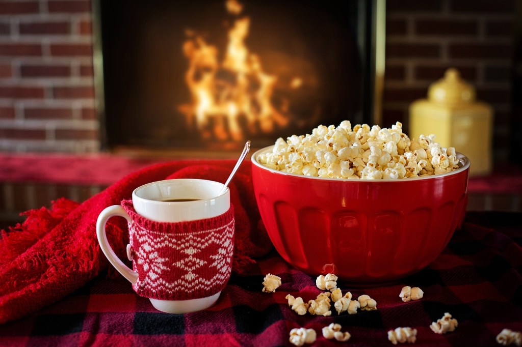 A cozy scene featuring a red bowl filled with popcorn next to a white mug with a knitted red sleeve, all set near a warm fireplace, with a festive red blanket draped nearby.
