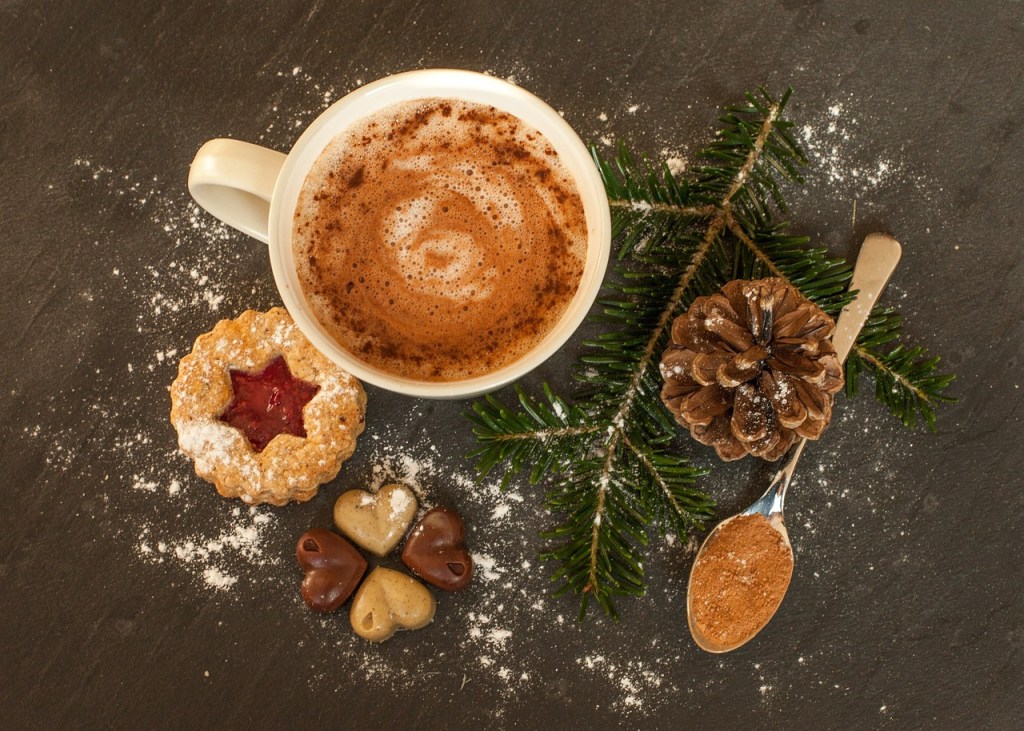 A warm cup of hot chocolate with a swirling pattern, accompanied by festive cookies, a pine cone, a spoon with cinnamon, and evergreen sprigs, set against a dark surface.
