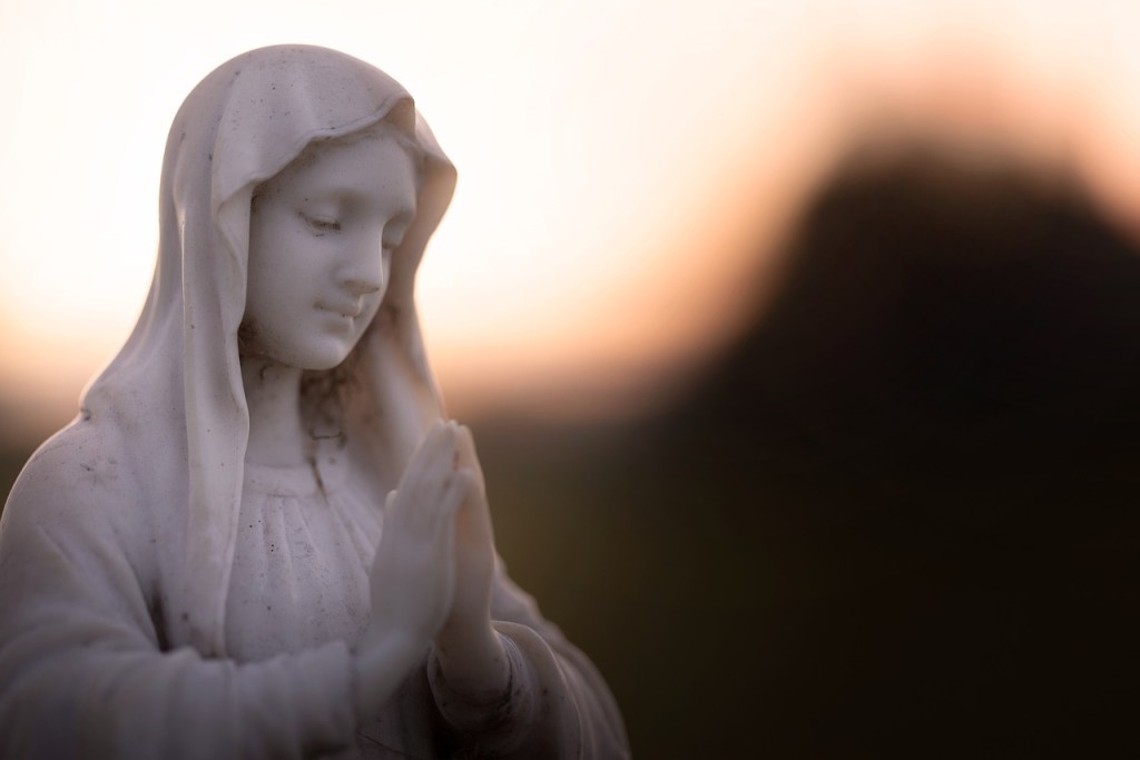 A serene statue of the Virgin Mary with clasped hands, symbolizing prayer and devotion, set against a soft-focus background of warm colors.