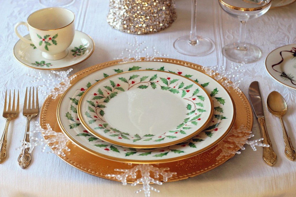 A beautifully arranged holiday table setting featuring decorative plates with holly designs, a teacup on a saucer, utensils, and crystal accents.
