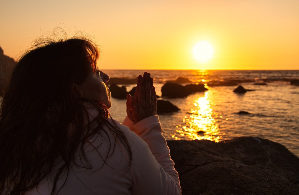 A person with long hair kneels in prayer at sunset by the ocean, with the sun reflecting on the water.