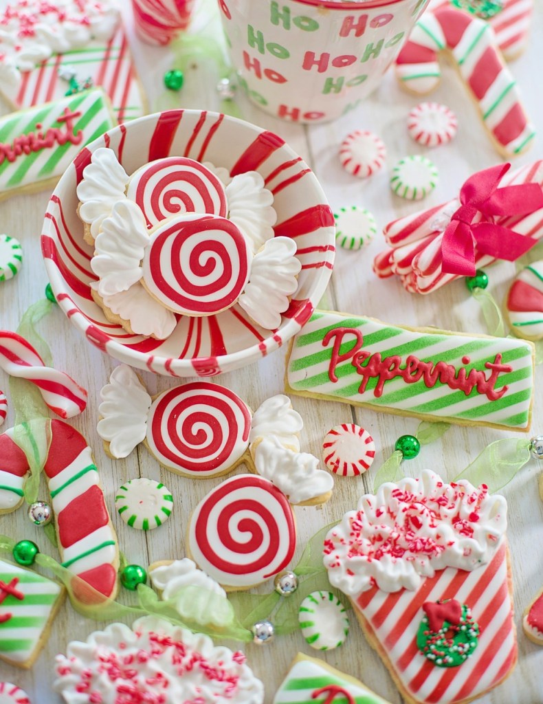 A festive arrangement of decorated Christmas cookies in various shapes and colors, including peppermint-themed cookies, candy canes, and holiday-themed sweets, all presented in a bowl and scattered across a light wooden surface.