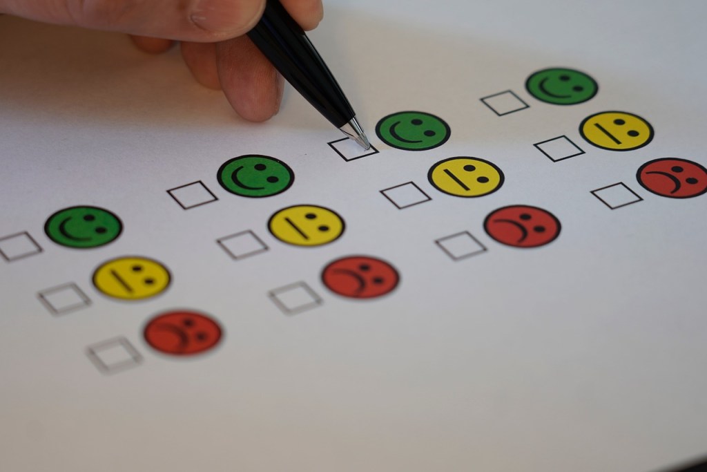 A hand holding a pen checks off options on a printed feedback form featuring colorful smiley faces representing different emotions.