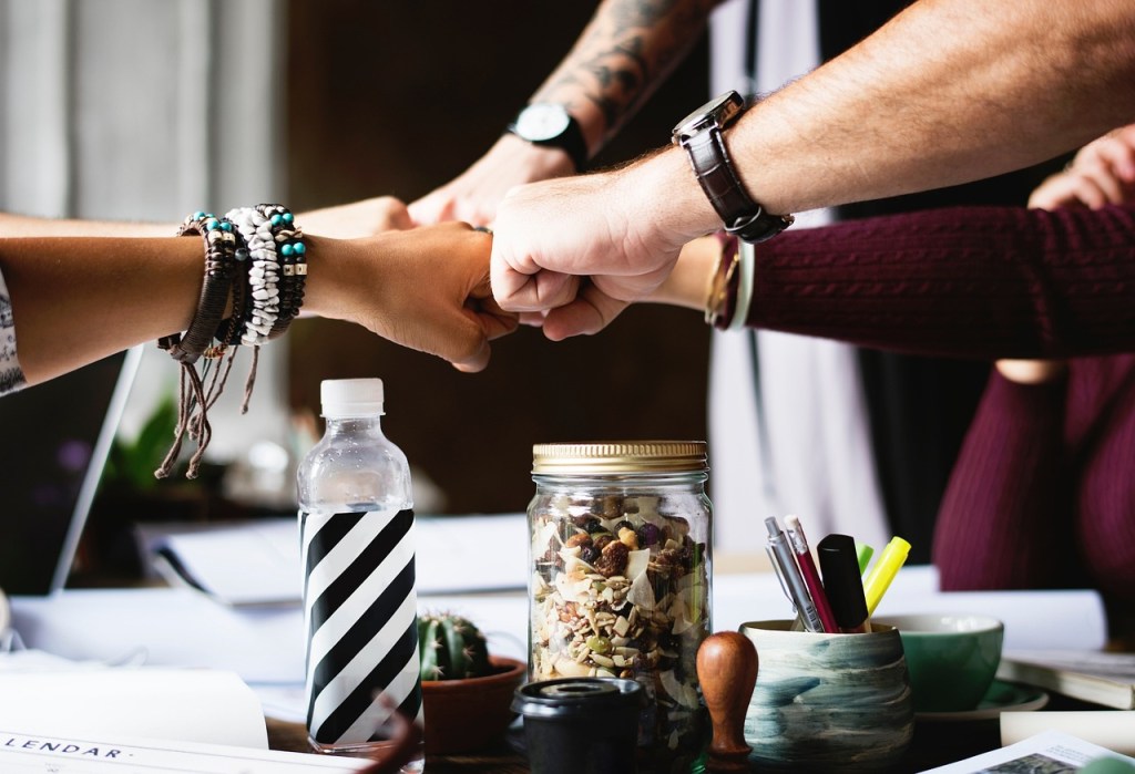 A group of diverse hands performing a fist bump over a table filled with various items, including a striped water bottle, a jar of snacks, and pens in a holder.