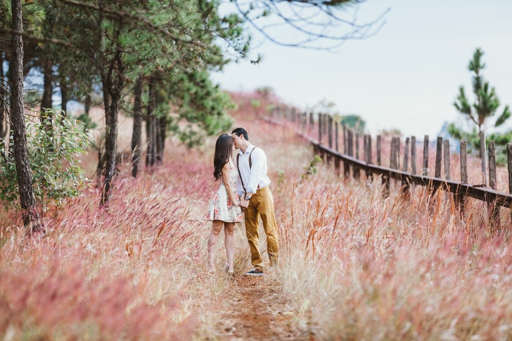 A couple stands closely together in a colorful grassy field surrounded by trees, sharing an intimate moment.
