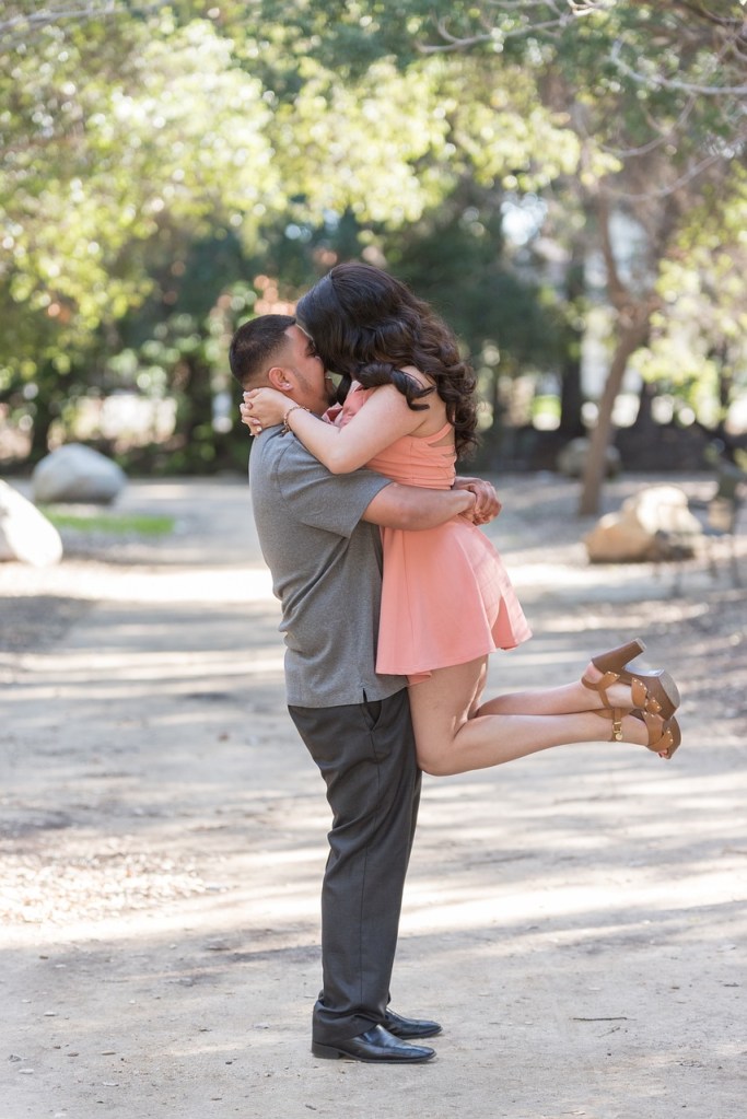 A couple sharing an intimate moment outdoors, with the man lifting the woman while they kiss, surrounded by trees and a path.