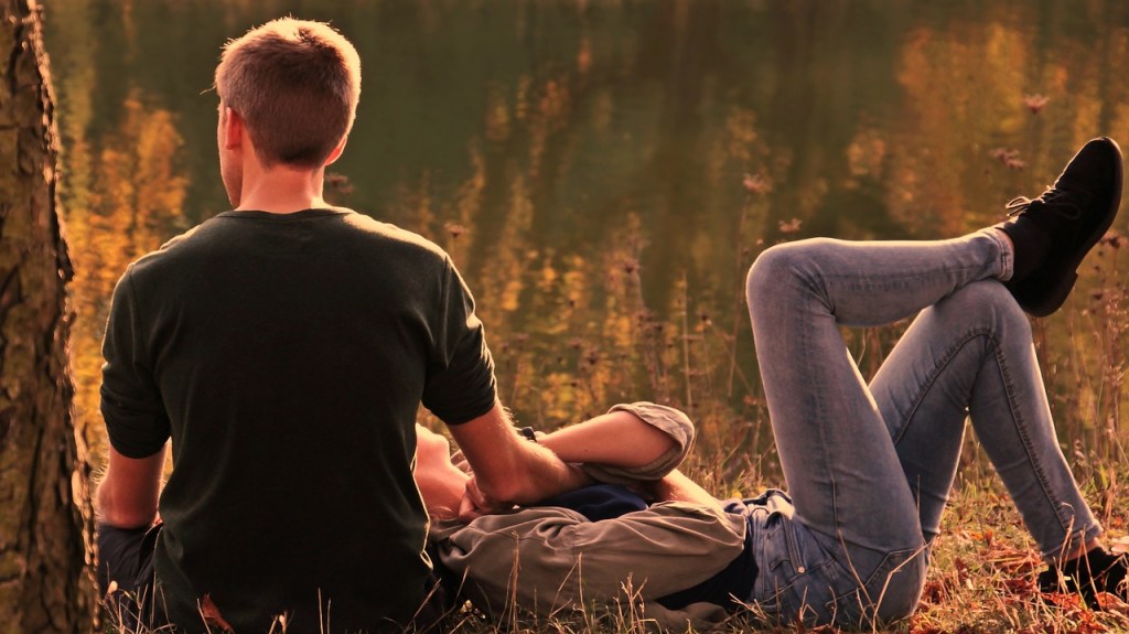 A young couple sitting together by a lake, sharing an affectionate moment with the woman resting her head on the man's shoulder.