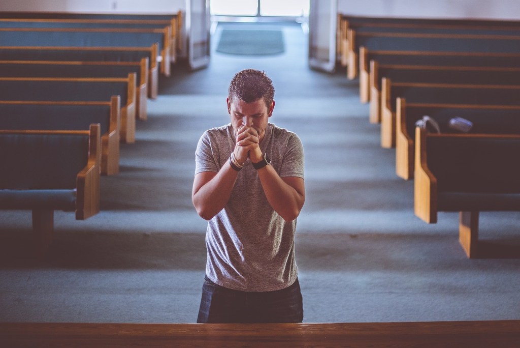 A man praying in a church, standing with his hands clasped together, facing an altar, with empty pews in the background.